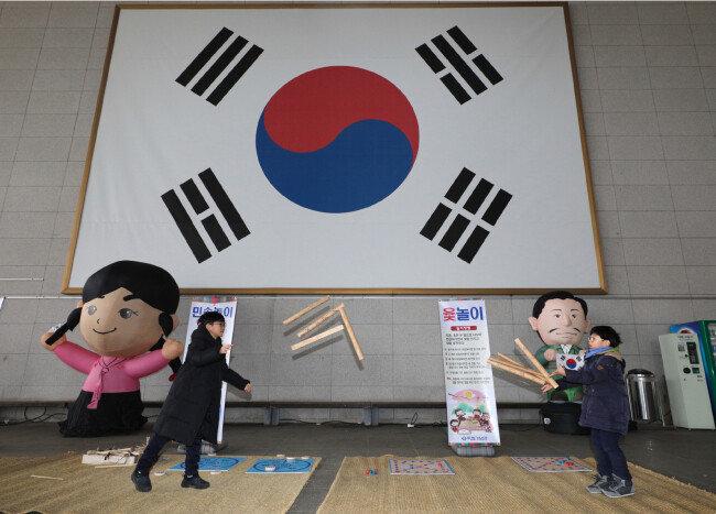 독립의 상징인 대형 태극기를 배경 삼아 두 어린이가 윷놀이를 하고 있다.
