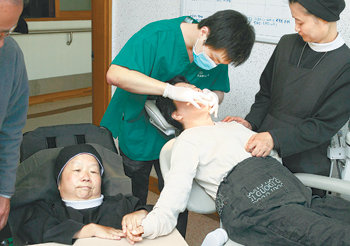 푸르메나눔치과 오승환 원장이 26일 경기 가평군 ‘성가정의 집’을 방문해 중증장애인 환자를 치료하고 있다. 치과 진료를 무서워하는 환자를 위해 윤석인 원장수녀(왼쪽)가 침대형 휠체어에 누워 환자의 손을 잡아주고 있다. 사진 제공 푸르메재단