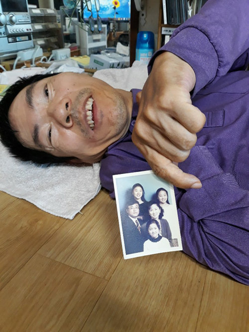 몸을 가누기 어려워 대부분 누워 지내는 이대우 씨가 만나고 싶다는 ‘제네트 김’의 가족사진을 보여주고 있다. 제네트 김은 사진 속 가운데 줄 오른쪽 여성이다. 이대우 씨 측 제공