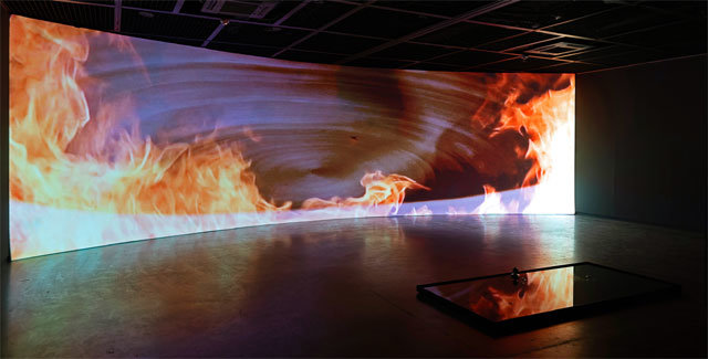 영상설치작품 ‘Beyond’. 검은 물 앞에 놓인 너비 11m, 높이 2.7m의 대형 스크린 속에서 불꽃에 휩싸인 도기 물레가 회전한다. 작가는 “죽음을 머금은 삶에 대해 관조할 계기를 마련하고 싶었다”고 했다. 성북구립미술관 제공