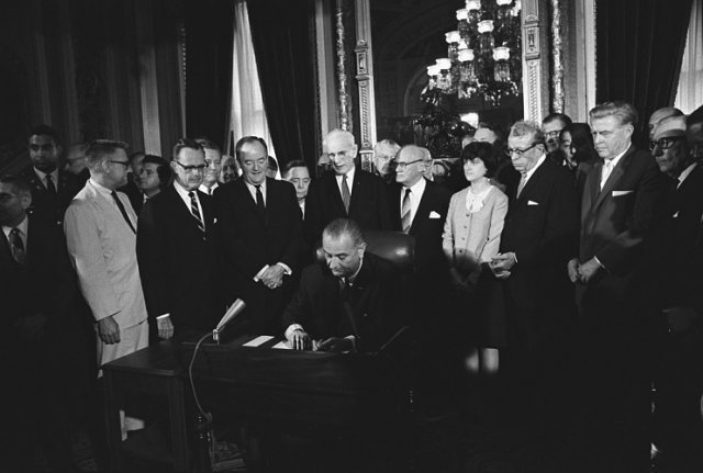 1965년 백악관에서 열린 투표권법 서명식에서 당시 18세이던 린든 존슨 대통령의 딸 루시 (오른쪽 4번째))가 아버지가 서명하는 모습을 지켜보고 있다. 린든 존슨 도서관 홈페이지