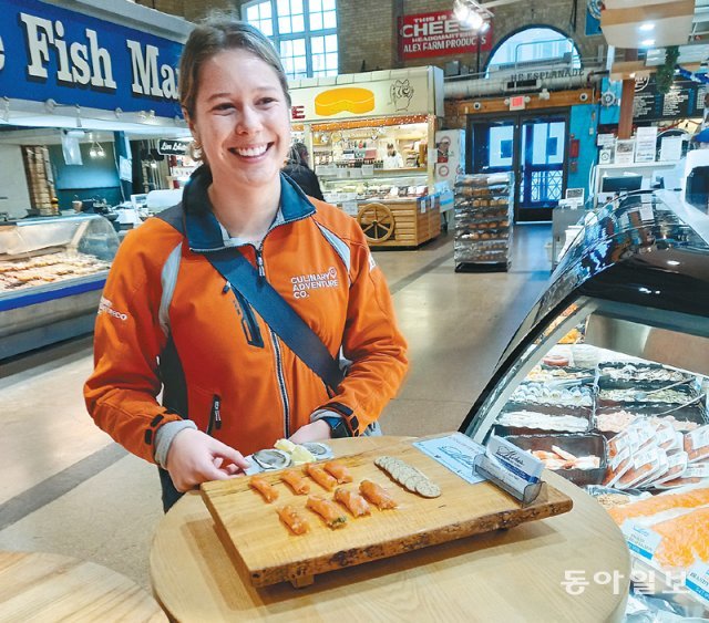 토론토의 전통시장 세인트로렌스마켓을 함께 둘러보며 캐나다의 식재료와 음식에 대해 설명해 주는 미식투어 프로그램이 있다. 안내자가 시장 내에서 구입할 수 있는 다양한 방식으로 훈제된 연어의 시식을 권하고 있다.