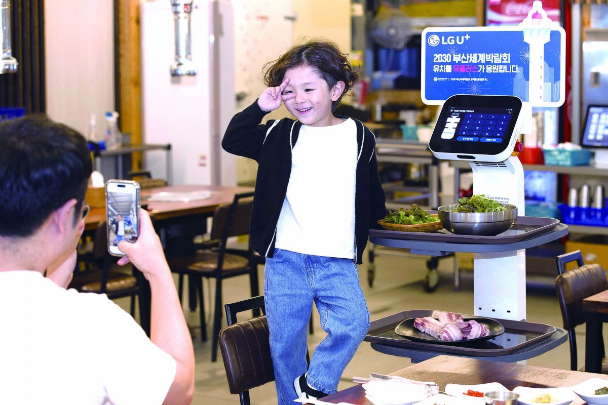 A customer of Dawn Charcoal Garden Busan Daeshin branch takes a commemorative photo with a U+ serving robot attached to a cheering panel for the Busan Expo.