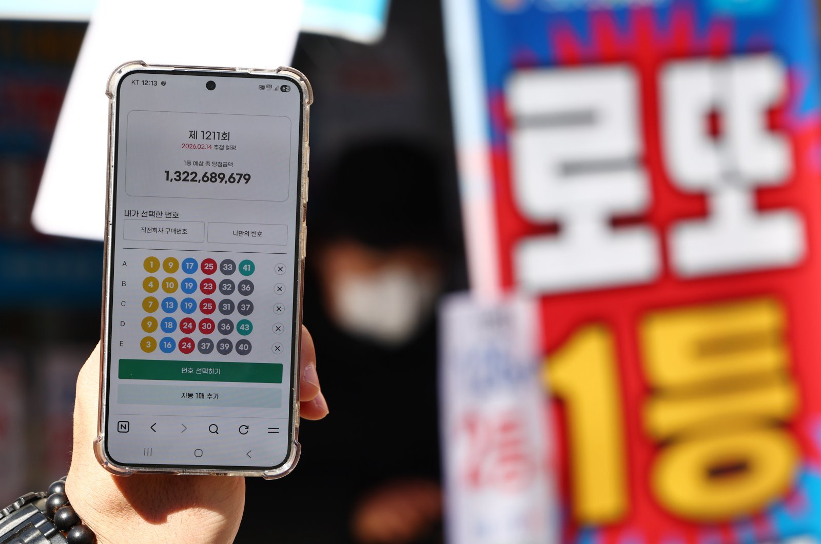 24년 만에 모바일 로또 판매가 시작된 9일 오전 서울 시내 한 복권 판매점 앞에서 휴대폰 화면에 나오는 로또 모바일 판매 페이지 뒤로 시민들이 로또를 구매하고 있다.  이용자는 모바일 동행복권 홈페이지에 가입한 뒤 예치금을 충전하면 오전 6시부터 자정까지 복권을 살 수 있다. 올해 상반기에는 평일에만 회차당 1인 5000원까지 살 수 있으며, PC 온라인 구매 금액과 합산해 계산된다. 토요일에는 모바일 구매가 막힌다. 전체 온라인 판매 규모도 전년도 로또 판매액의 5% 이내로 묶여, 한도에 도달하면 판매가 자동 중단된다. 2026.02.09. [서울=뉴시스]