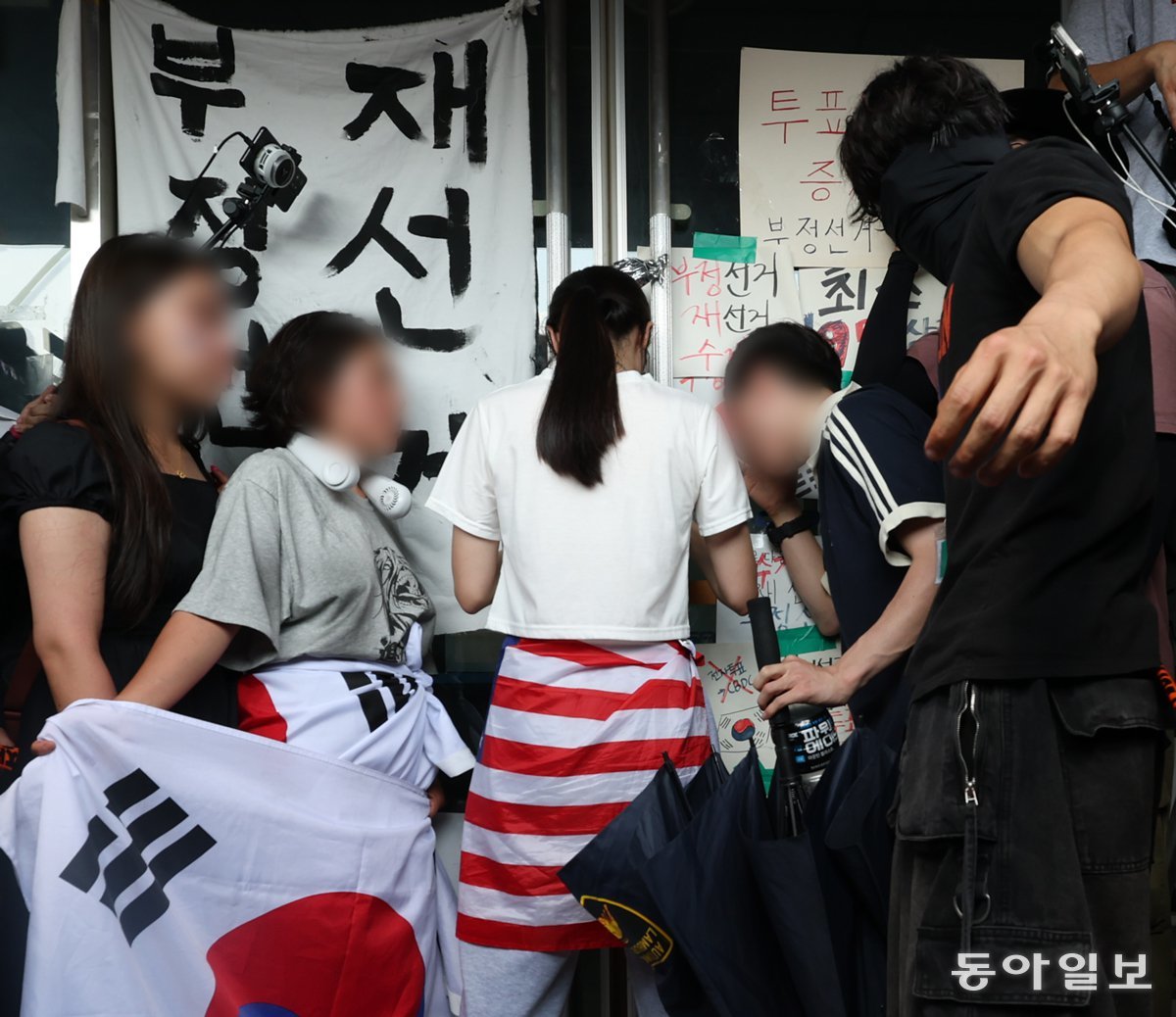 19일 서울 송파구 올림픽공원 핸드볼경기장 앞에서 6·3 지방선거 투표용지 부족 사태를 규탄하는 집회가 열린 가운데, 흰옷을 입고 성조기를 허리에 두른 여성(가운데)이 경기장 문을 잡고 버티고 있다. 이날 경기장에 입주한 대한체육회 산하 체육단체 관계자는 집회 참가자들과 ‘생중계 카메라 대동’ 등의 조건에 합의하고 건물에 진입하려 했지만 이 여성이 비켜주지 않아 끝내 들어가지 못했다. 변영욱 기자 cut@donga.com