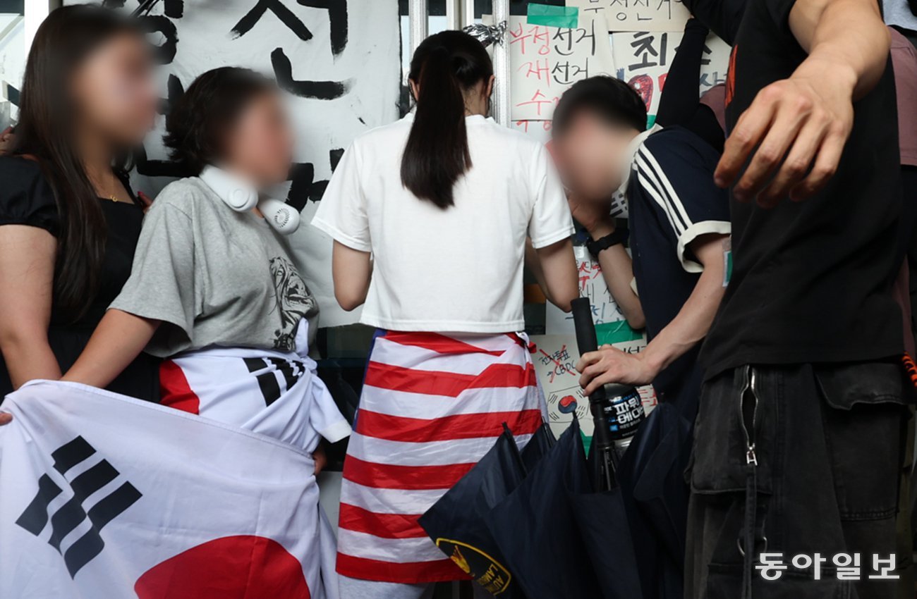 19일 서울 송파구 올림픽공원 핸드볼경기장 앞에서 6·3 지방선거 투표용지 부족 사태를 규탄하는 집회가 열린 가운데, 흰옷을 입고 성조기를 허리에 두른 여성(가운데)이 경기장 문을 잡고 버티고 있다. 이날 경기장에 입주한 대한체육회 산하 체육단체 관계자는 집회 참가자들과 ‘생중계 카메라 대동’ 등의 조건에 합의하고 건물에 진입하려 했지만 이 여성이 비켜주지 않아 끝내 들어가지 못했다. 변영욱 기자 cut@donga.com