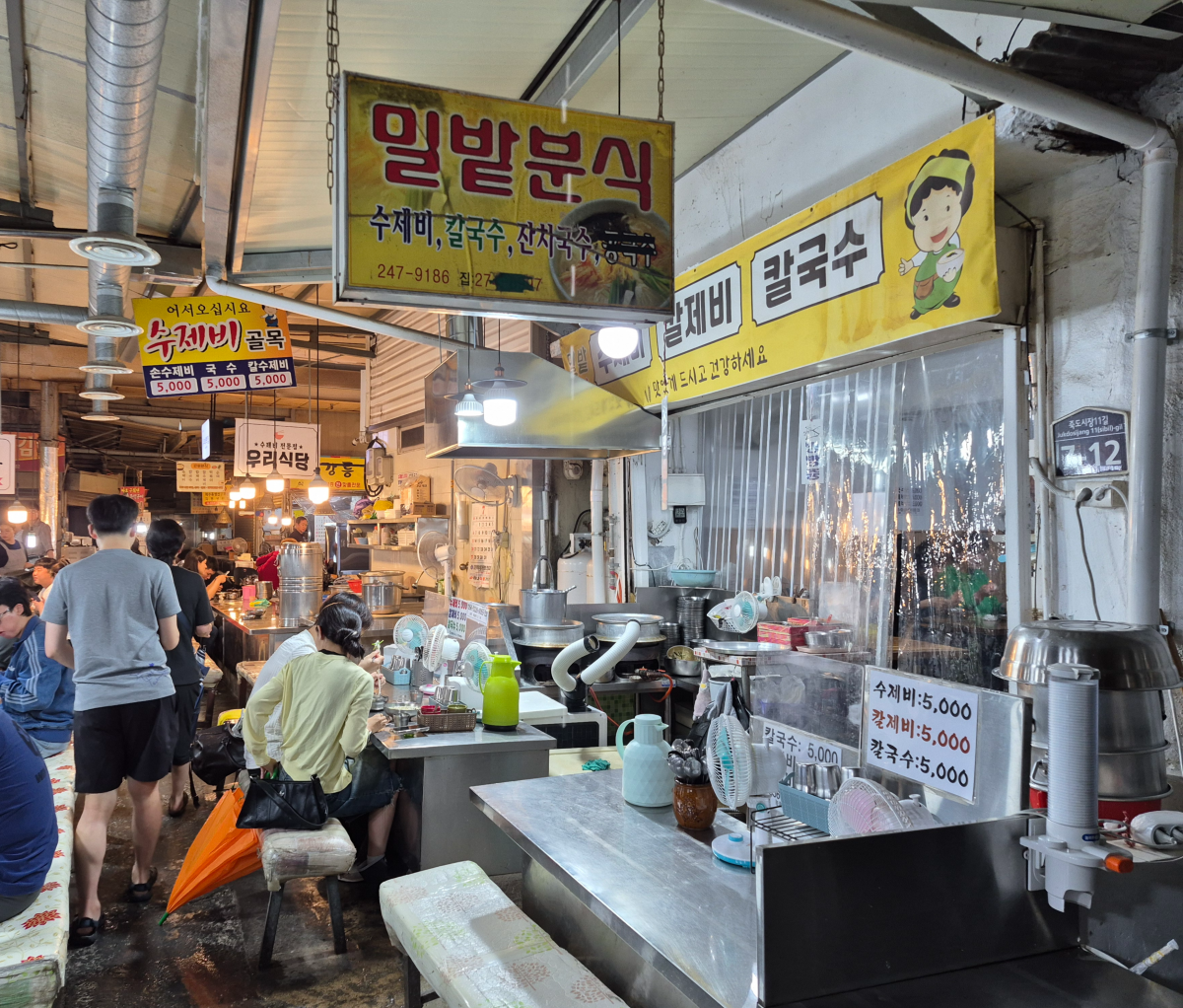 비 오는 날의 포항 죽도시장 수제비골목. 긴 의자에 앉아 수제비를 먹고 있는 사람들을 볼 수 있다.